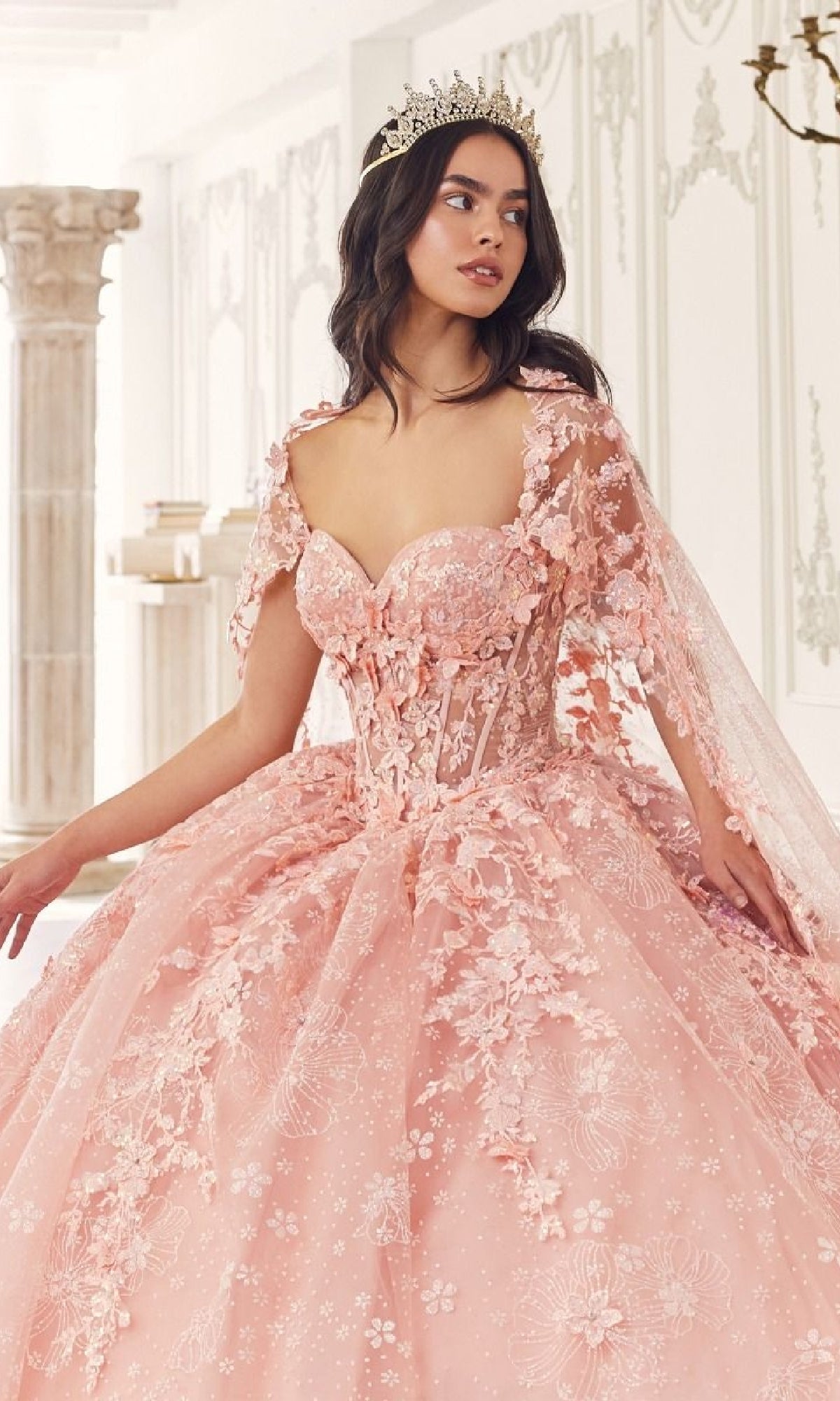 A woman wears the Quinceanera Dress 15719 by Ladivine, a light pink floral-embroidered gown, and a crown while posing in an elegant classical room with white walls and columns.